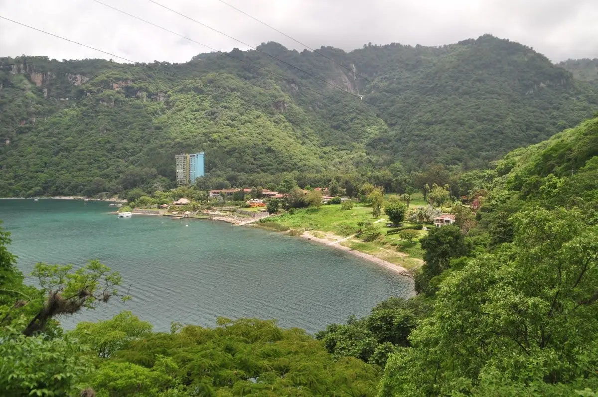 View of Atitlán Nature Reserve Bay west of Panajachel, with Lake Atitlán curving along the shore, dense green hillsides, and a tall lakeside building beneath overcast skies.