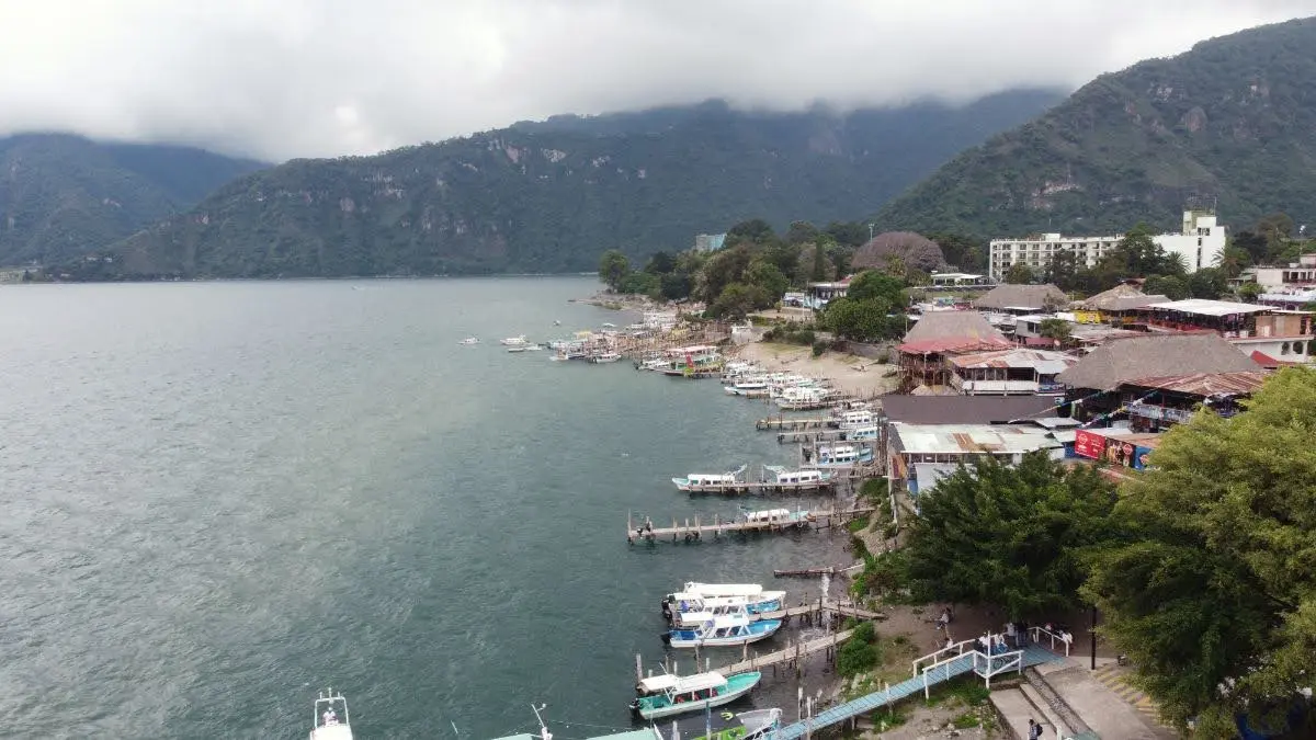 Wide view of Lake Atitlán with Panajachel’s shoreline, lakeside docks, and green mountains partly covered by low clouds, seen from the water.