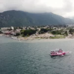 Wide view of Lake Atitlán with Panajachel’s shoreline in the background, green mountains under low clouds, and a small pink passenger boat crossing the water.