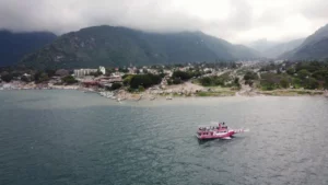 Wide view of Lake Atitlán with Panajachel’s shoreline in the background, green mountains under low clouds, and a small pink passenger boat crossing the water.