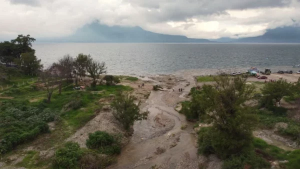 San Francisco River flowing into Lake Atitlán at Panajachel, with a shallow riverbed, green vegetation, and cloud-covered mountains in the distance.