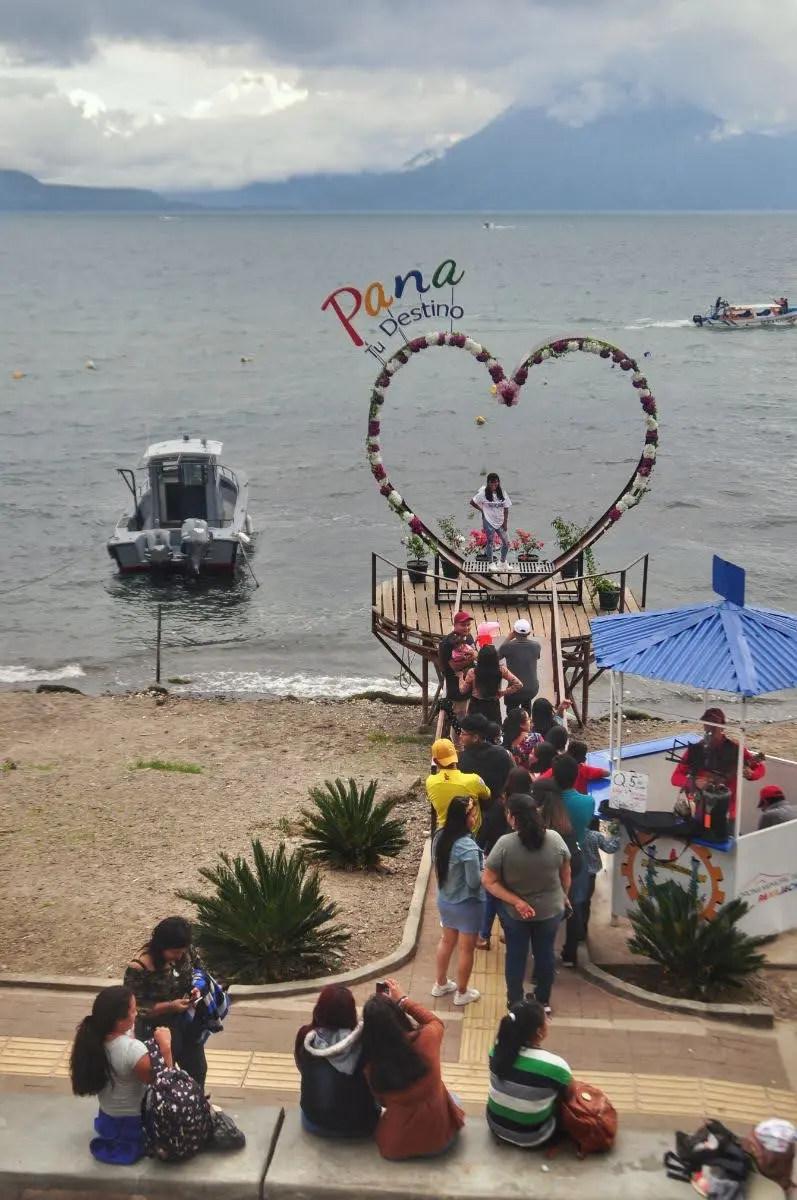 People lining up at the heart-shaped “Pana” photo spot on the shore of Lake Atitlán in Panajachel, with the lake, boats, and cloud-covered mountains in the background.