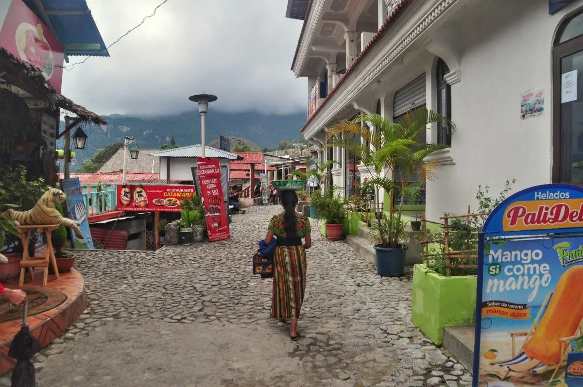Woman walking along the malecon in Panajachel, passing small shops and street vendors, with colorful storefronts and cloud-covered mountains in the background.