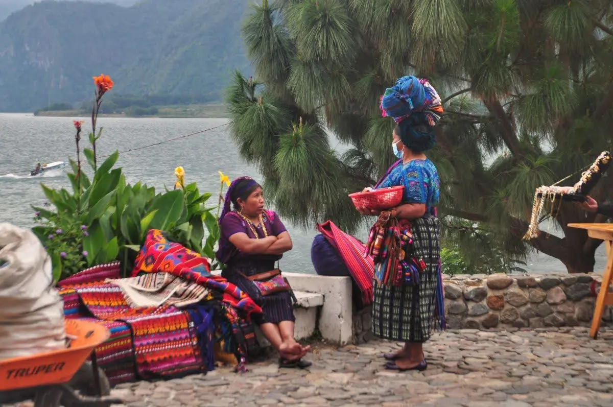 Indigenous women selling textiles by Lake Atitlán in Panajachel, with woven fabrics displayed on a stone wall and the lake and mountains in the background.