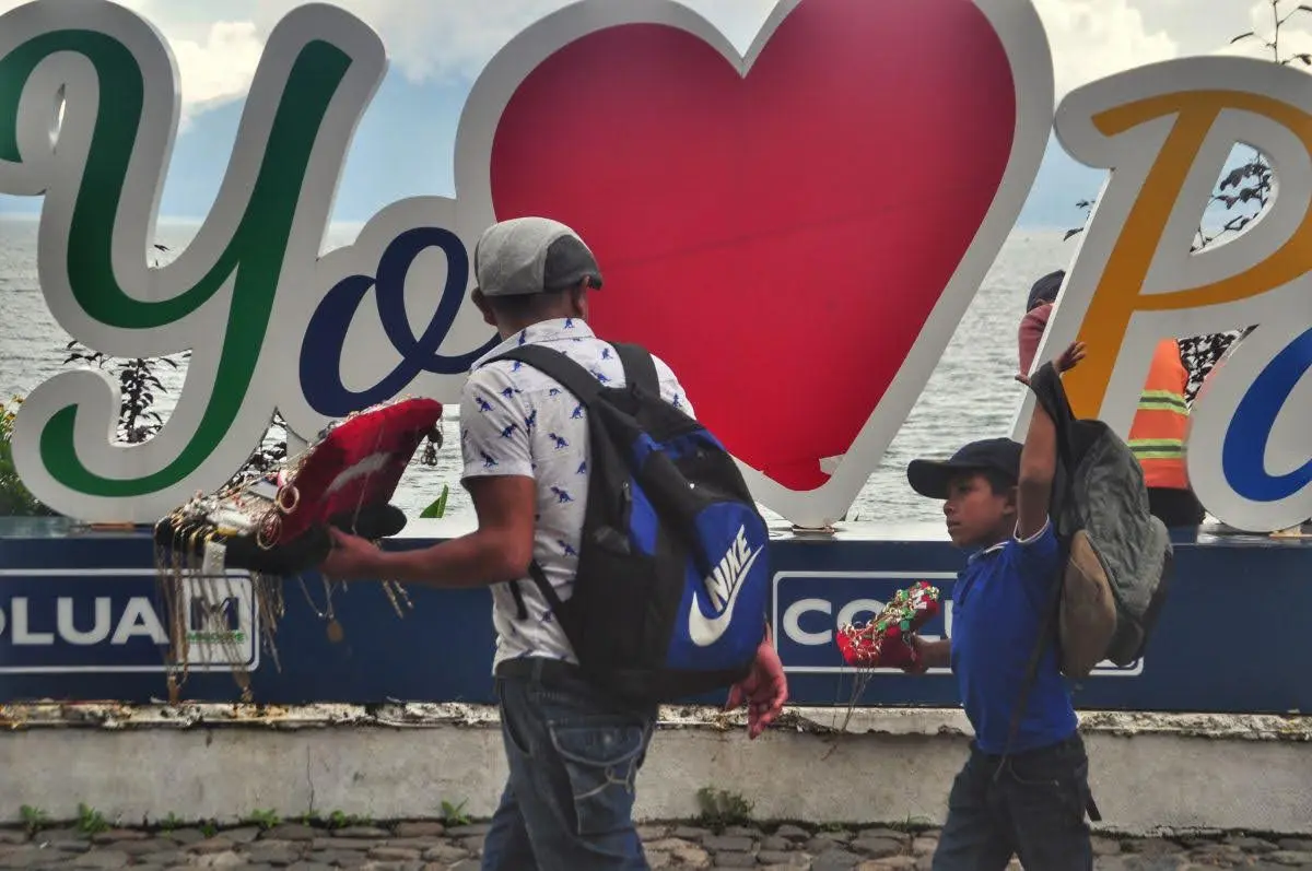 Father and son walking along Panajachel’s waterfront, carrying goods in front of the “Yo Amo Panajachel” sign by Lake Atitlán.