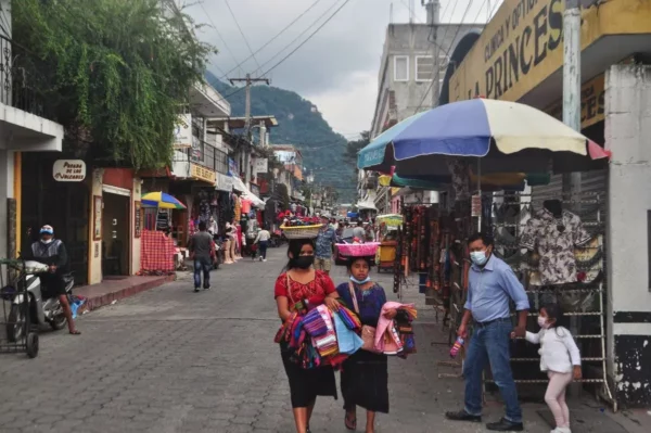 Two Indigenous women walking along Santander street in Panajachel, carrying woven textiles, with market stalls, pedestrians, and hillside buildings in the background.