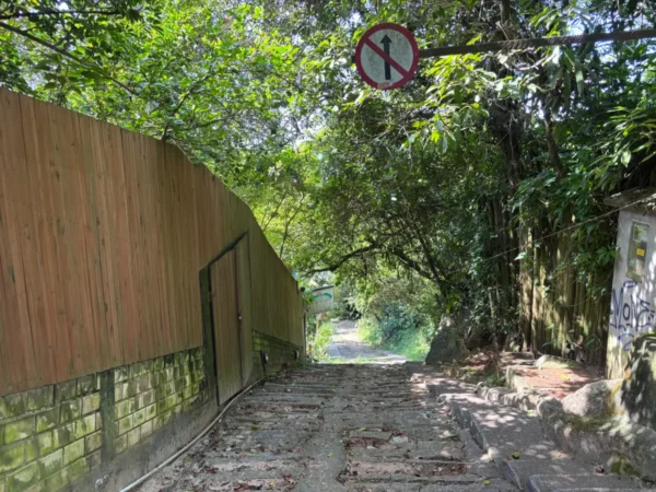 Overgrown path leading to Praia do Rosa, with dense greenery, a wooden fence, and a no-entry sign in Santa Catarina, Brazil.