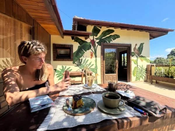 Relaxing at a café table with coffee, pastel-colored walls adorned with tropical plants, and a sunny outdoor setting in Praia do Rosa, Santa Catarina, Brazil.