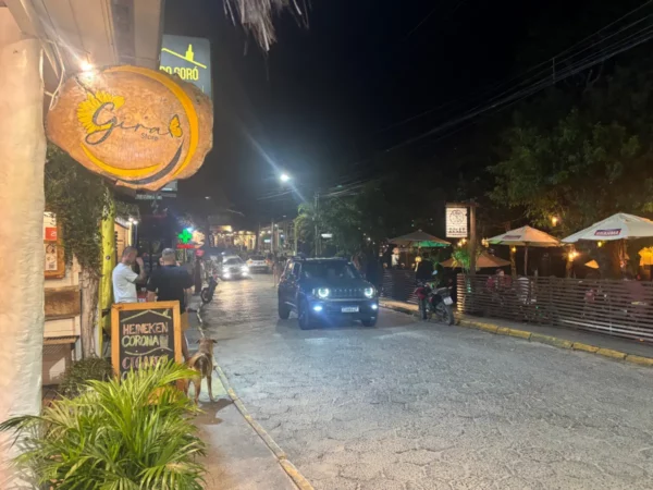 Bustling street scene in Praia do Rosa, Santa Catarina, Brazil, featuring shops, dining, and a car illuminated by streetlights.