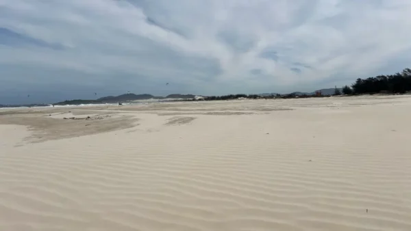 Waves gently roll onto the sandy shore at Praia do Rosa, surrounded by lush greenery and a cloudy blue sky, Santa Catarina, Brazil.