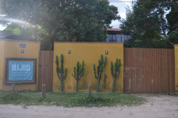 Bright yellow walls with cacti complement the rustic wooden fence at Praia do Rosa, Santa Catarina, Brazil. Lush greenery surrounds the area.