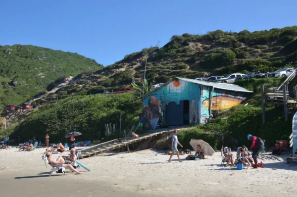 Colorful beach shack sits on a sandy shore, with sunbathers and lush hills behind at Praia do Rosa, Santa Catarina, Brazil.