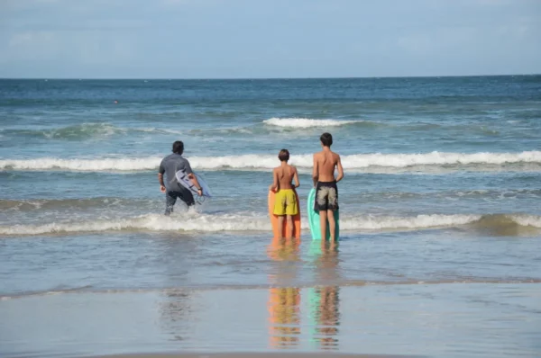 Three children wade into the gentle waves at Praia do Rosa, Santa Catarina, Brazil, with bright surfboards reflecting on the water.