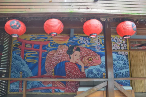 Colorful red lanterns hang above a mural depicting a woman and a large fish, set against a vibrant blue and gold background in Praia do Rosa, Santa Catarina, Brazil.