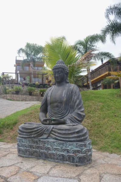 Buddha statue surrounded by greenery in Praia do Rosa, Santa Catarina, Brazil, with residential buildings in the background.