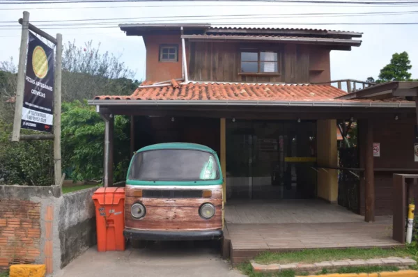 Rustic wooden building with a brown tiled roof, featuring an old van and a bright orange mailbox at Praia do Rosa, Santa Catarina, Brazil.