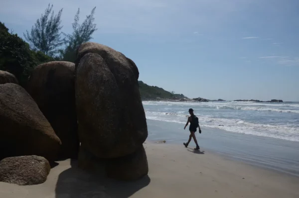 Large rocks line the sandy beach at Praia do Rosa, with a lone figure walking along the shoreline in Santa Catarina, Brazil.