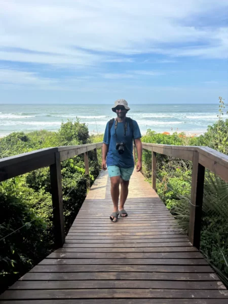A person walks along a wooden boardwalk surrounded by lush greenery, leading to Praia do Rosa beach in Santa Catarina, Brazil.