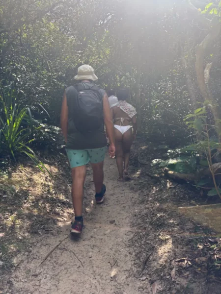 Hikers navigate a sandy path through lush greenery near Praia do Rosa in Santa Catarina, Brazil. Sunlight filters through the trees.