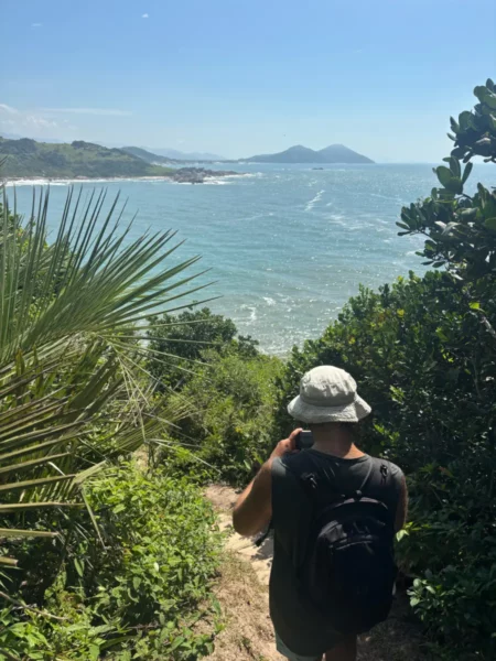 Walking through lush greenery, a person descends towards the sparkling waters of Praia do Rosa, Santa Catarina, Brazil.