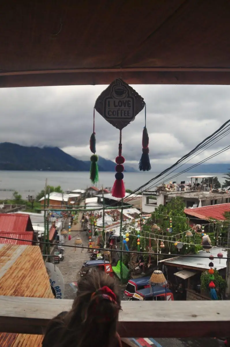 View from a terrace café in San Juan La Laguna overlooking the main street and Lake Atitlán, with pedestrians below, colorful decorations, and mountains across the water.
