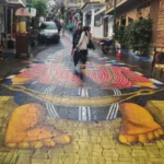 Pedestrians walking along a narrow street in San Juan La Laguna, Guatemala, with a colorful mural painted across the pavement depicting traditional Maya imagery.