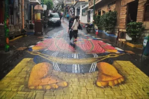 Pedestrians walking along a narrow street in San Juan La Laguna, Guatemala, with a colorful mural painted across the pavement depicting traditional Maya imagery.