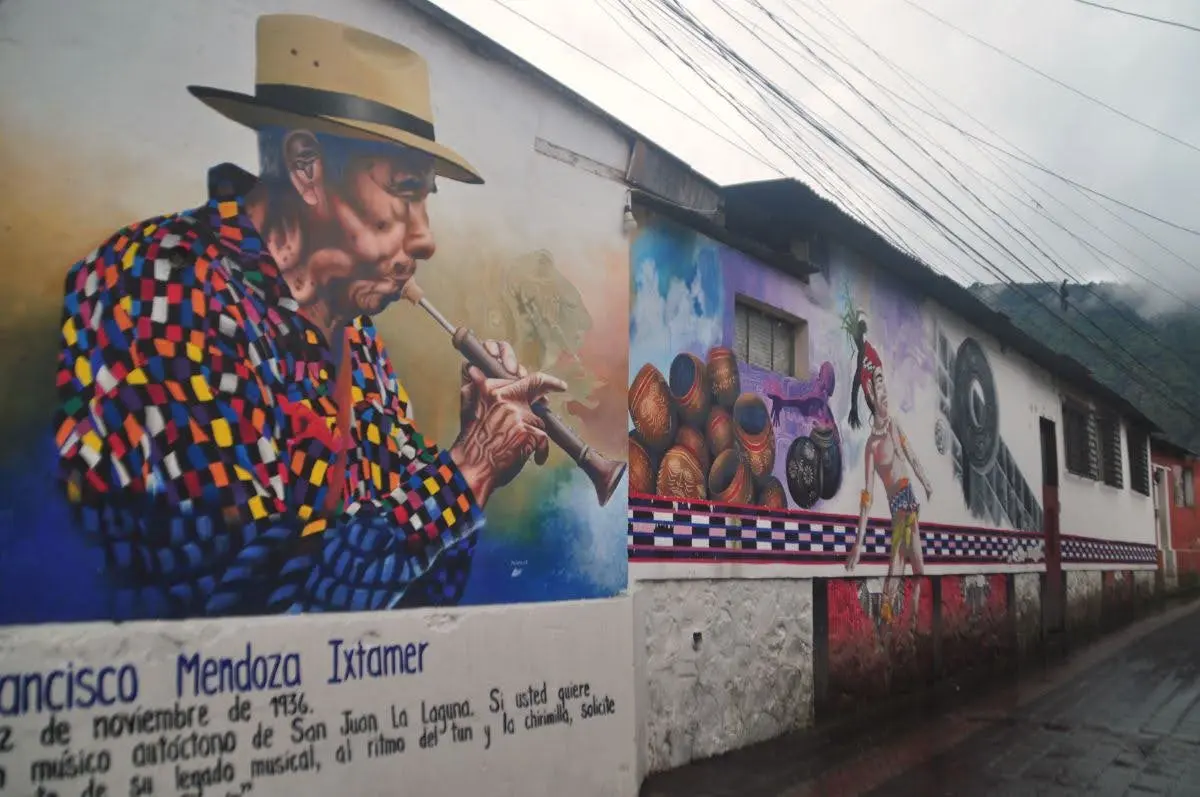 Street mural in San Juan La Laguna depicting a local Maya man playing a flute, painted along a narrow village street in Guatemala.