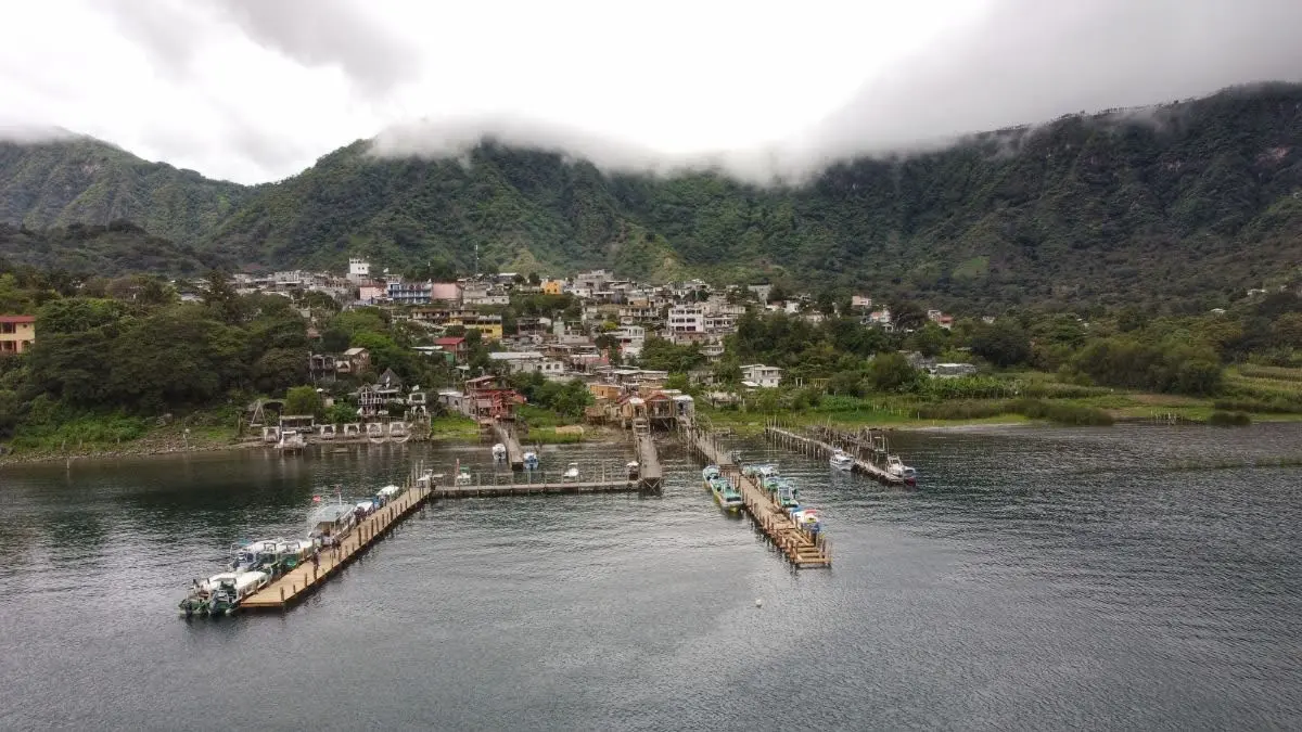 drone view of San Juan La Laguna from Lake Atitlán, showing the village waterfront with two wooden piers, boats at the dock, and green hills rising behind the town.