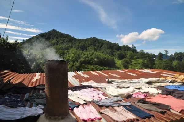 Smoking chimney rising from a corrugated metal rooftop in Santa Clara La Laguna, Guatemala, with clothes laid out to dry and forested highland hills in the background.