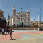 White colonial church facing a colorful village plaza in Santa Clara La Laguna, Guatemala, with locals gathered in the foreground and mountains under a clear sky.