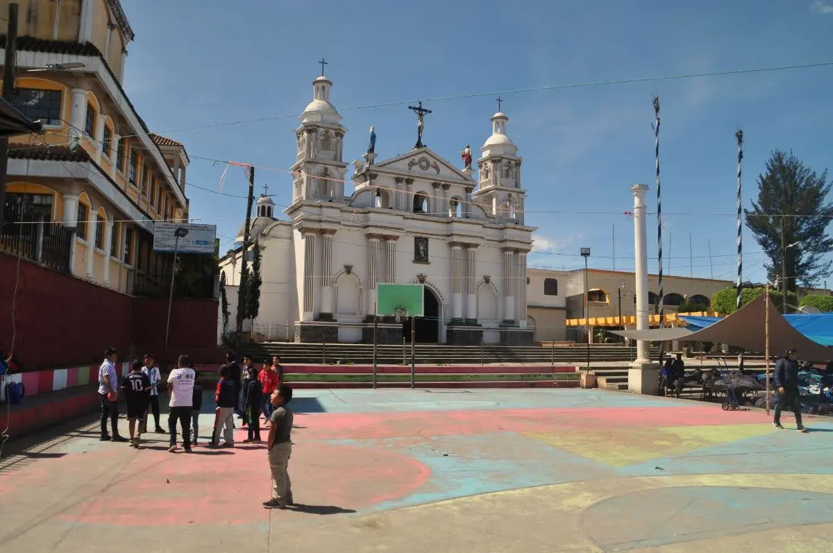 White colonial church facing a colorful village plaza in Santa Clara La Laguna, Guatemala, with locals gathered in the foreground and mountains under a clear sky.