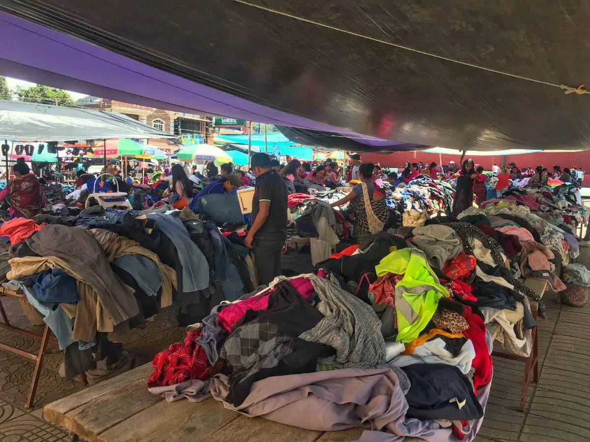 Crowded Sunday market in Santa Clara La Laguna, Guatemala, with piles of second-hand clothes spread across wooden tables under a large tarpaulin and locals browsing between stalls.
