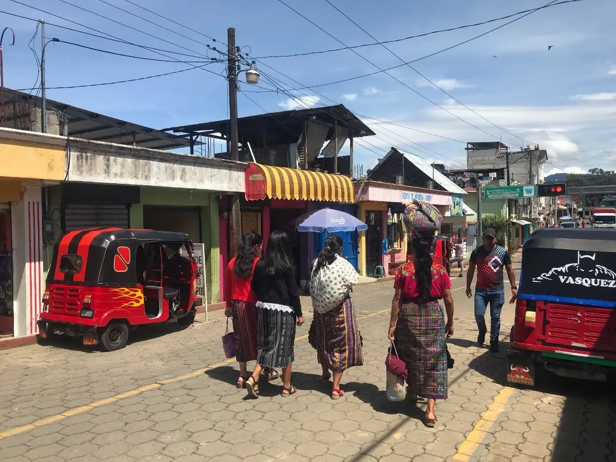 Women in traditional Maya dress walking along a street in Santa Clara La Laguna, Guatemala, with tuk-tuks, small shops, and market stalls lining the road.