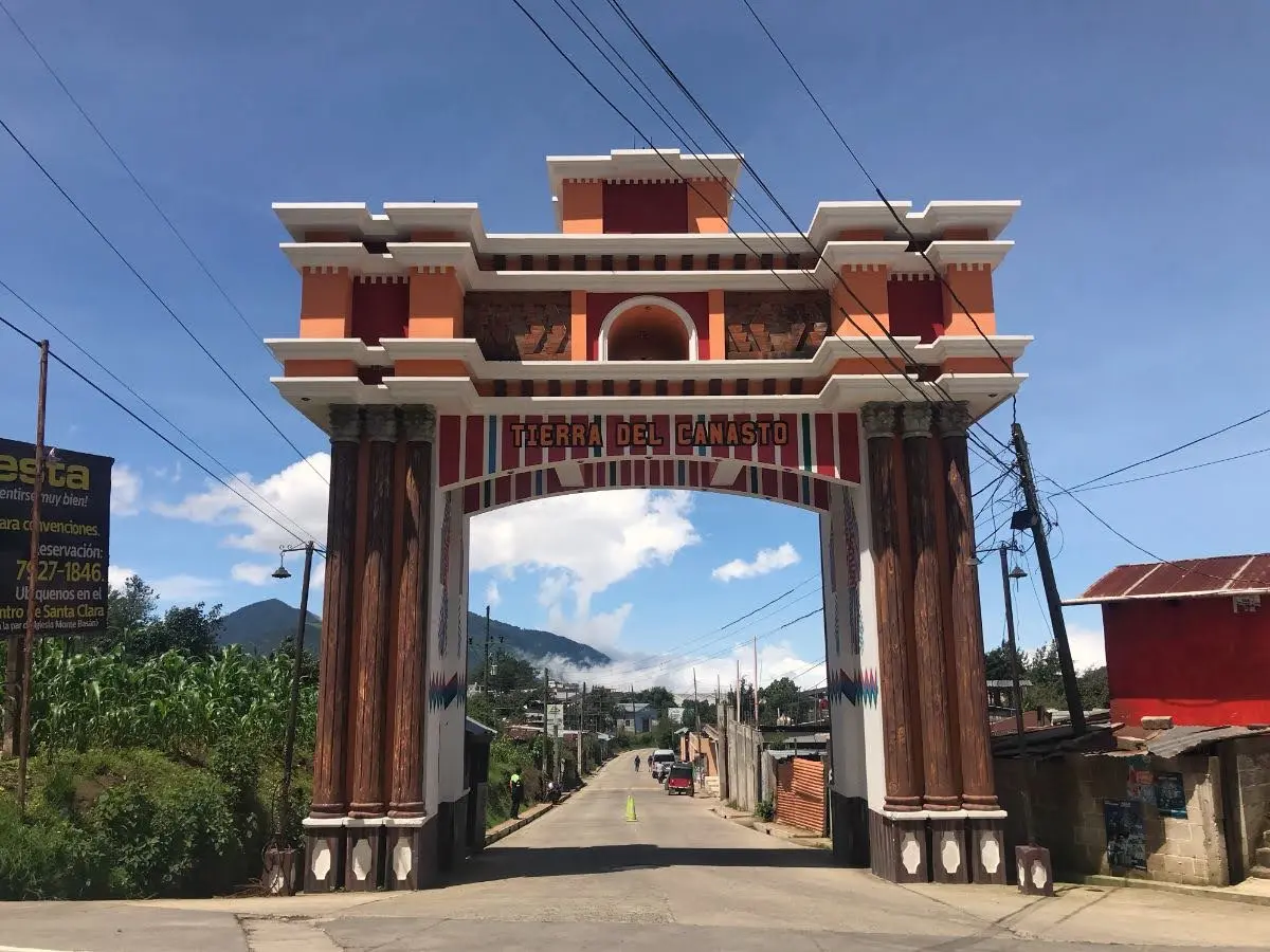 Ornate arched entrance gate of Santa Clara La Laguna, Guatemala, spanning the main road into town, with low houses, power lines, and hills visible beyond.