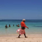 Vibrant waters of West Bay in Roatan, Honduras, with individuals swimming and a woman strolling along the sandy beach.