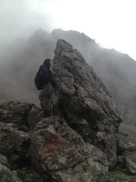 A hiker climbs a large rock formation on Xerovouni, surrounded by misty mountains and a dramatic cloudy sky.