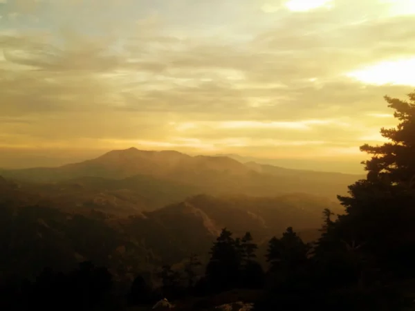 Golden sunset over the mountains at Xerovouni, with soft clouds and silhouettes of trees in the foreground.