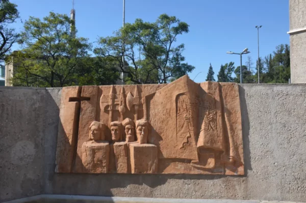 Relief sculpture featuring figures and a cross, set against a textured wall in Córdoba, Argentina, surrounded by trees.