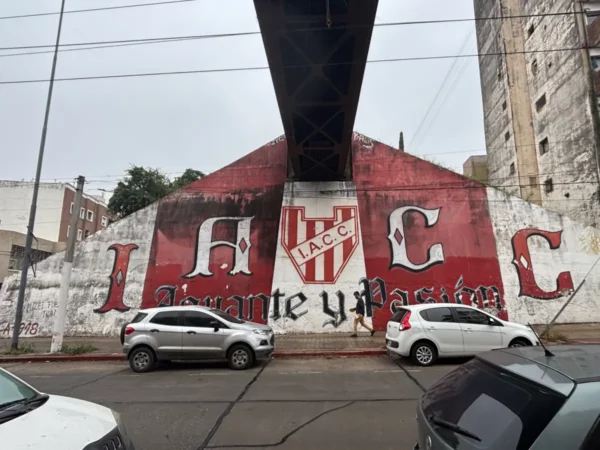 Colorful mural featuring the letters IACC and a shield, set against a concrete backdrop in Córdoba, Argentina. Cars are parked nearby.