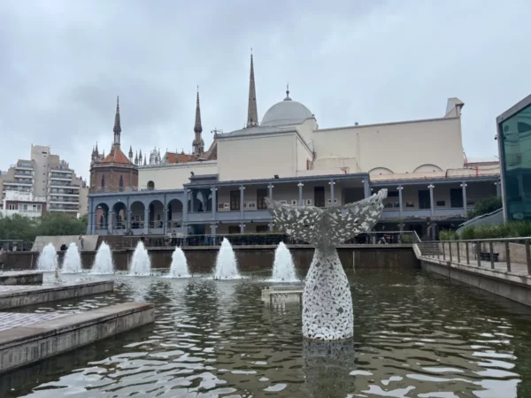 Fountain and decorative whale tail sculpture in front of historic buildings in Córdoba, Argentina, under a cloudy sky.