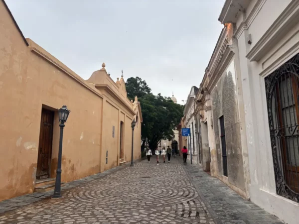 Historic cobblestone street with colorful colonial buildings in Cordoba, Argentina, lined by lush greenery and local pedestrians.