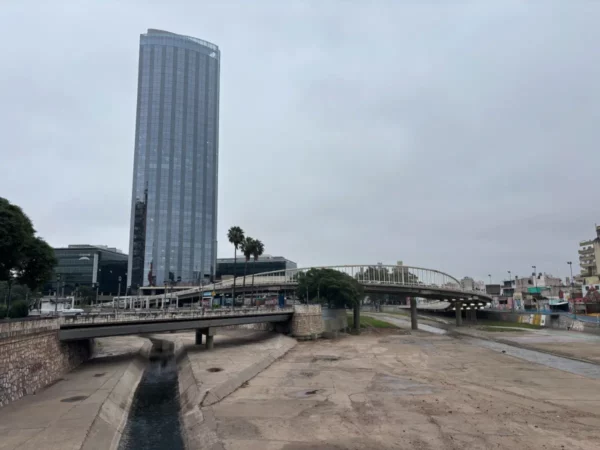 Modern high-rise buildings tower over the dry riverbed in Córdoba, Argentina, contrasting with palm trees along the waterfront.