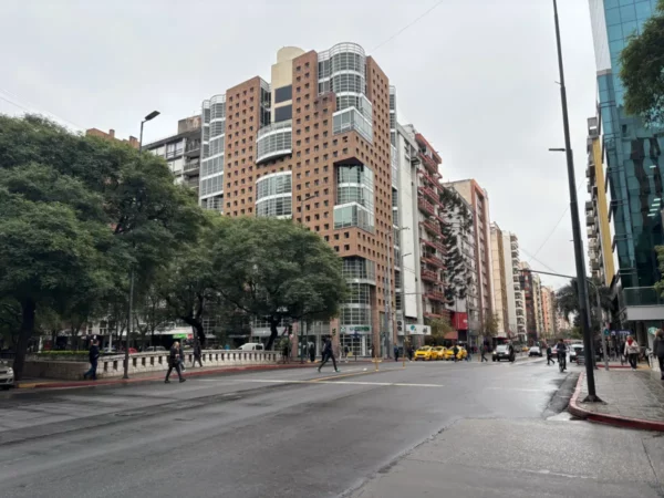 Modern buildings line a bustling street in Cordoba, Argentina, with trees providing shade and pedestrians crossing the road.
