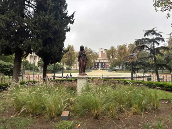 Statue surrounded by lush greenery at Plaza San Martín in Córdoba, Argentina, with city buildings visible in the background.