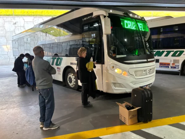 Travelers wait by a bus with a green destination sign in Córdoba, Argentina, showcasing urban transport and casual travel scenes.