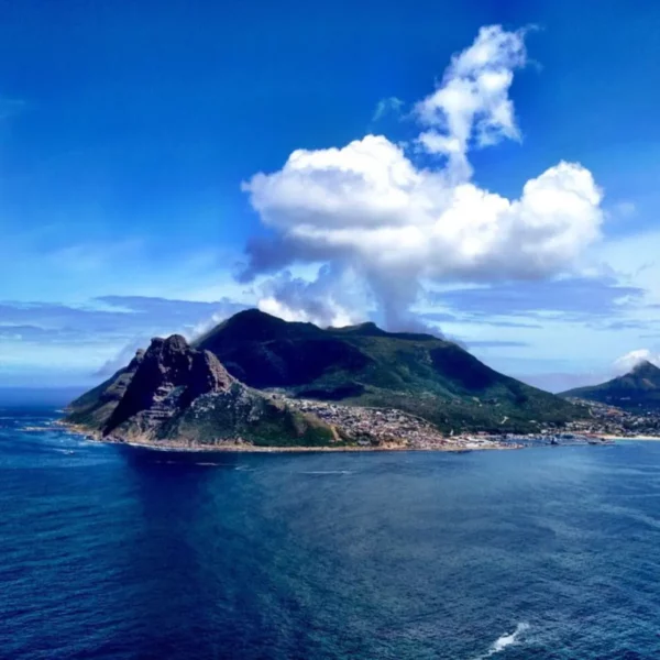 Stunning view of Hout Bay with rocky cliffs, vibrant blue waters, and fluffy clouds over the mountains in the background.