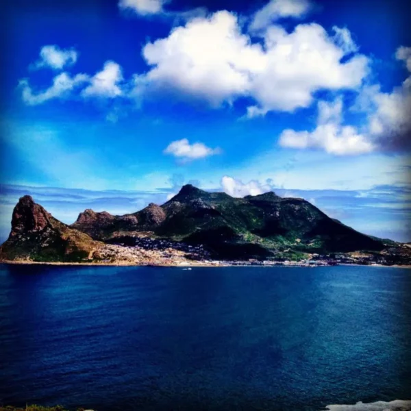 Hout Bay's stunning coastline features a vibrant blue ocean and dramatic green mountains under a bright sky with fluffy clouds.