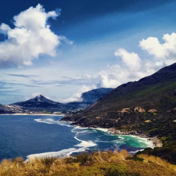 Stunning coastal view of Hout Bay with rolling waves, lush green hills, and fluffy clouds in a vibrant blue sky.