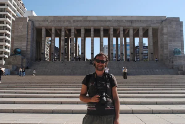 Man holding a camera stands on the steps of a grand, columned monument in Rosario, Santa Fe, Argentina, under a clear blue sky.
